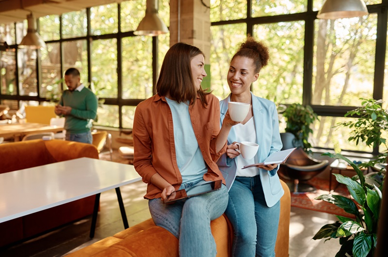Two successful young business woman enjoying pleasant conversation about startup project looking at digital tablet screen standing in eco friendly coworking space