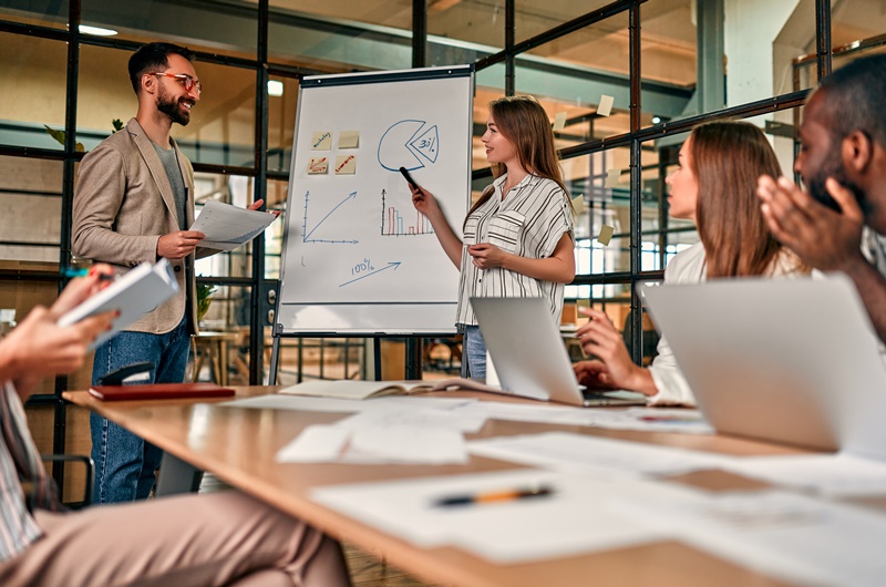 A young group of business people are discussing a new business plan on a blackboard, working on laptops while sitting in a modern office.